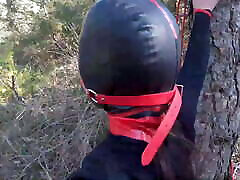 Tied up to a tree, outdoors in gloapi sex clothes, ball gagged